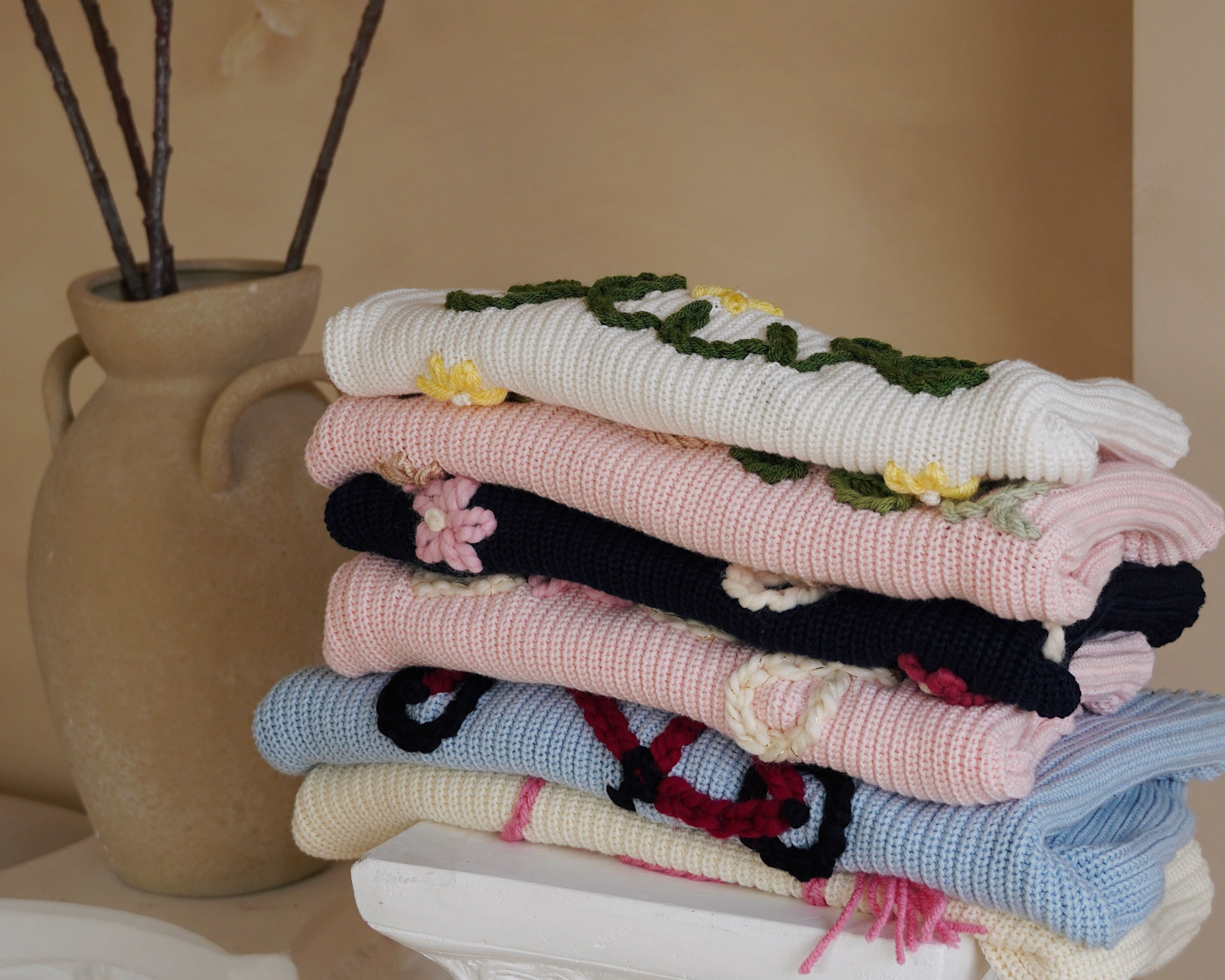 Stack of colorful knitted blankets on a decorative pedestal with a vase in the background.