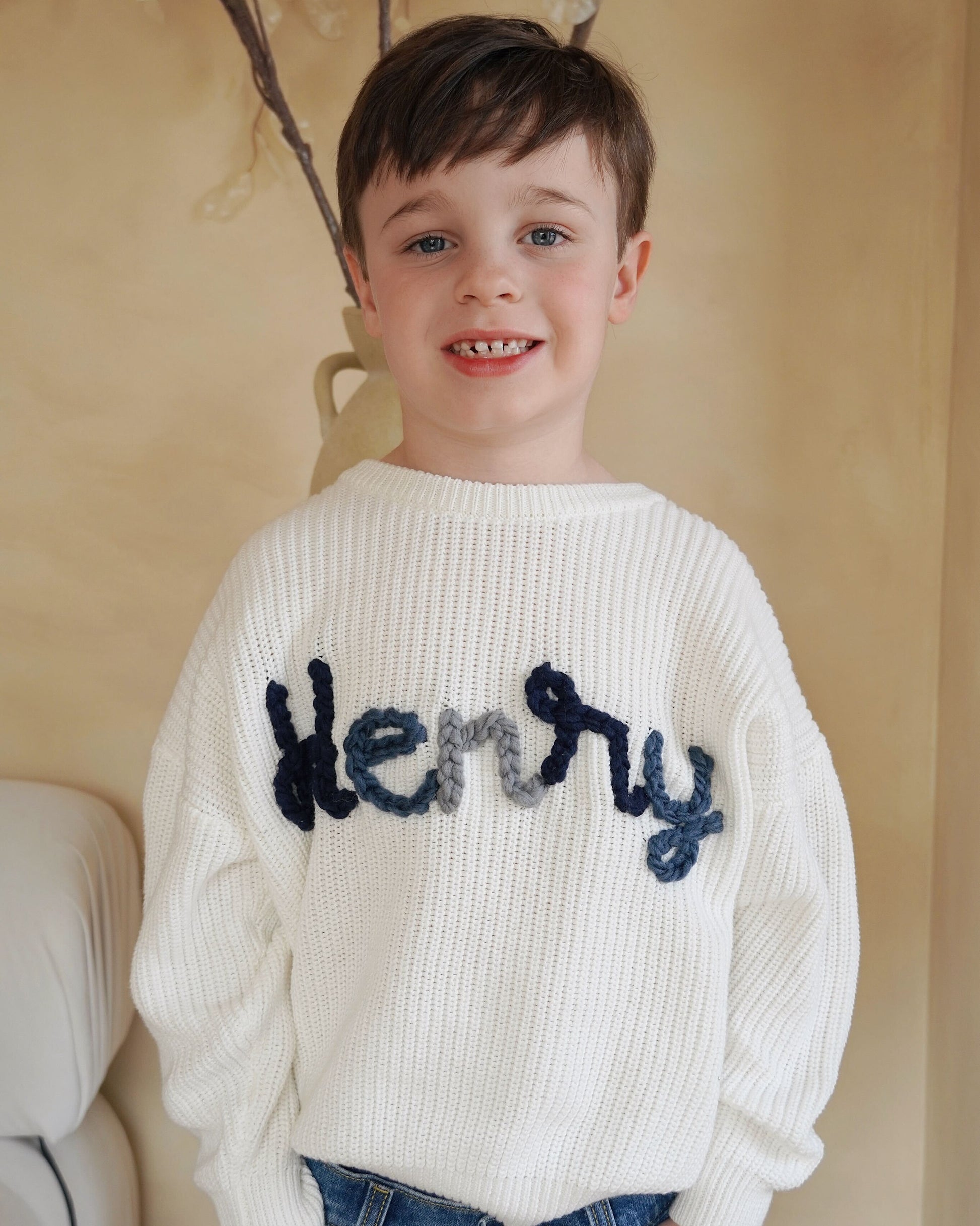 Child wearing a white sweater with 'Henry' printed on it, standing against a beige wall.