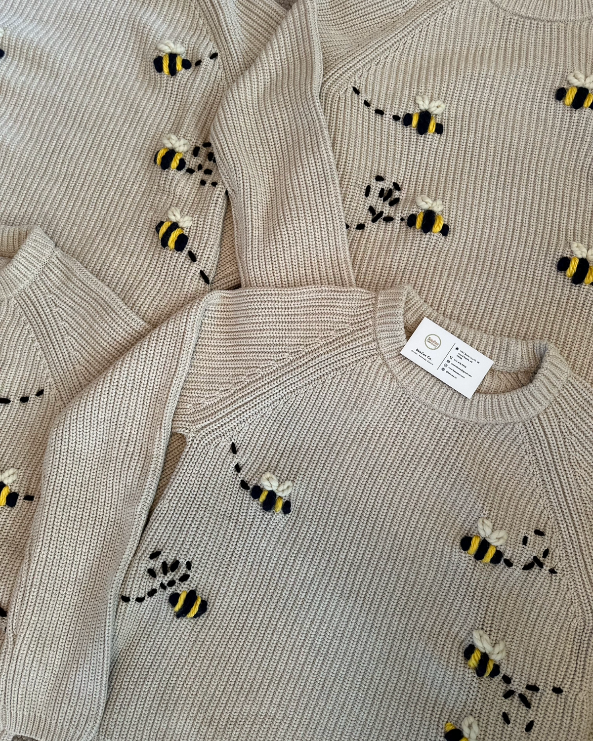 A close-up of a ribbed knit sweater in a light beige or oatmeal color, decorated with small embroidered bumblebees in black and yellow. Multiple views of the same sweater are shown overlapping.