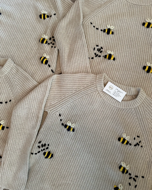 A close-up of a ribbed knit sweater in a light beige or oatmeal color, decorated with small embroidered bumblebees in black and yellow. Multiple views of the same sweater are shown overlapping.
