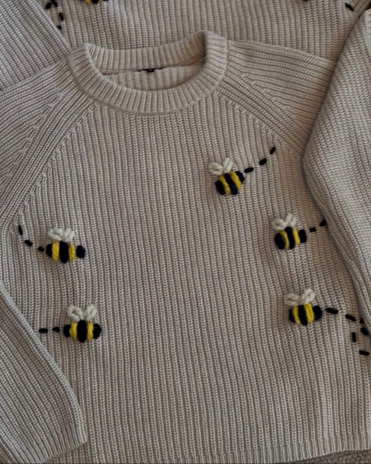 A close-up of a ribbed knit sweater in a light oatmeal beige color, decorated with small hand-knit bumblebee motifs in yellow, black, and white.