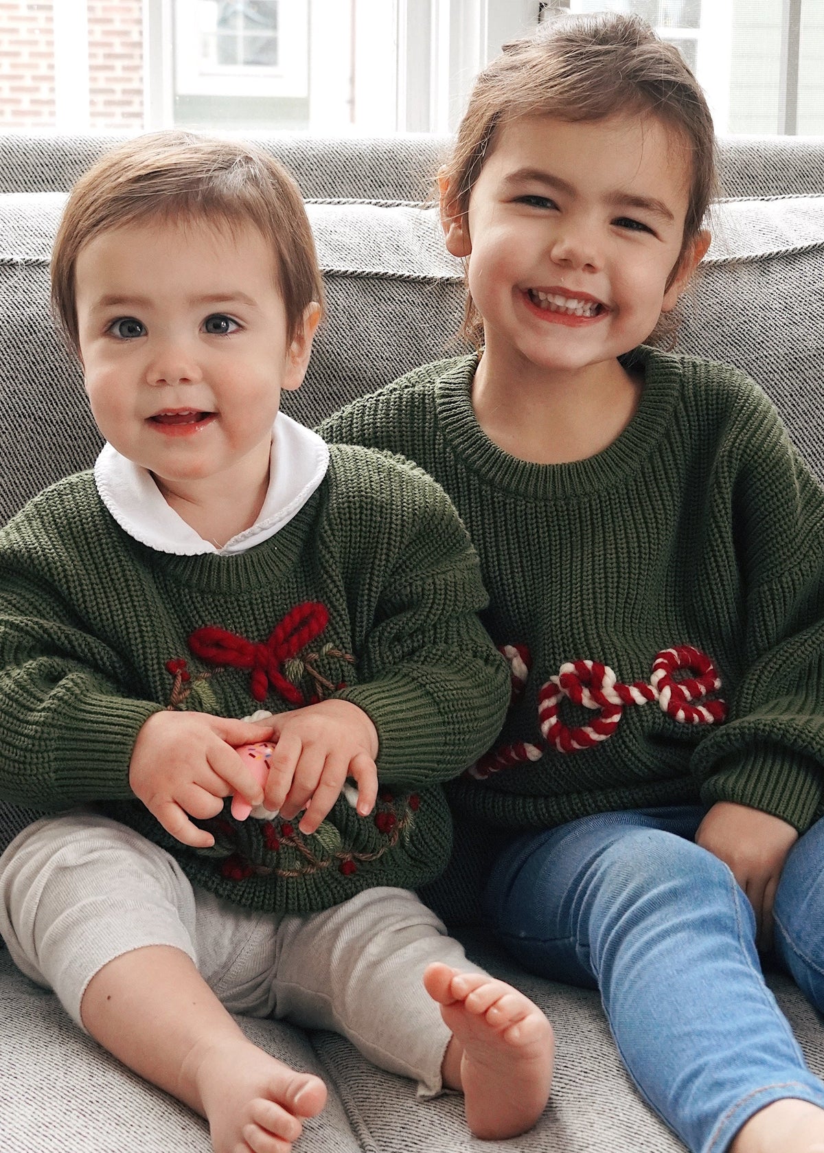 Two young children sitting on a gray couch indoors, wearing matching dark green knit sweaters with red and white holiday floral embroidery. The child on the left also wears a white collared shirt and light pants, while the child on the right wears blue pants.