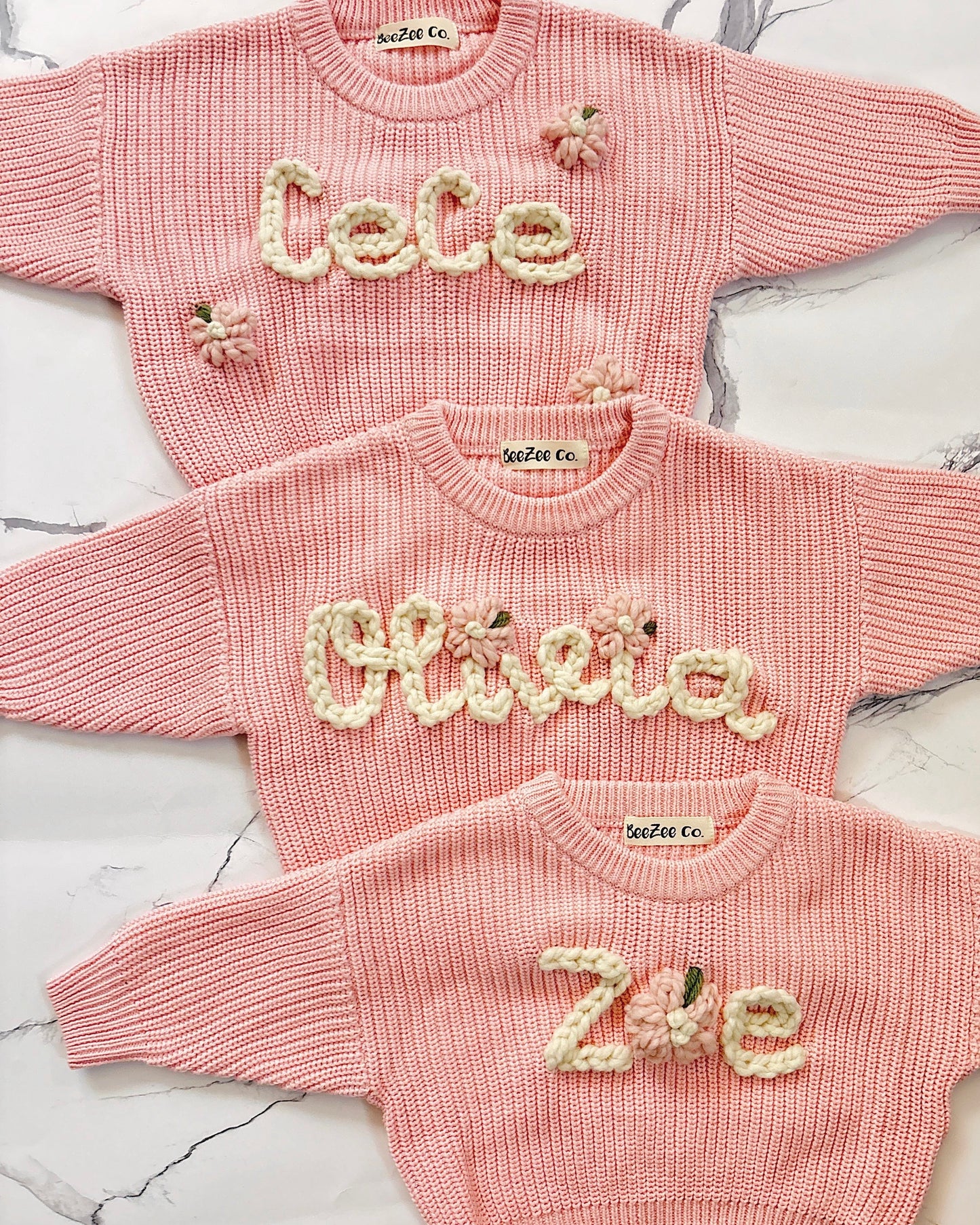 Three pink knitted baby outfits with embroidered names on a marble surface